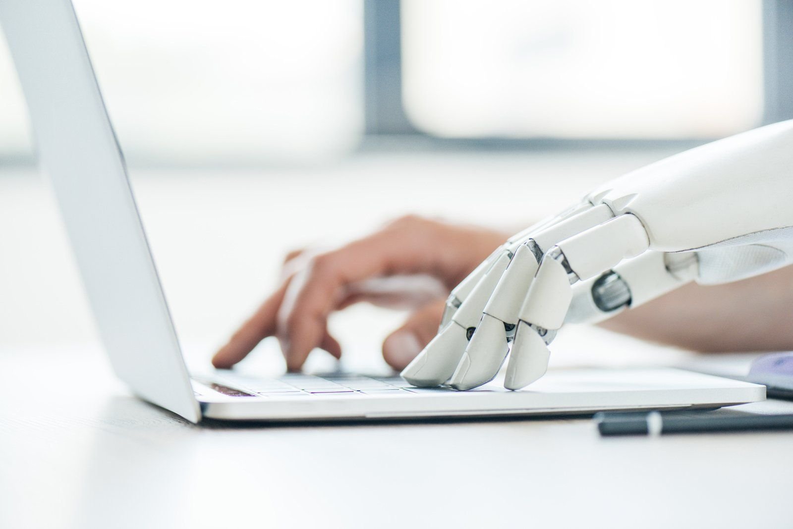 A robotic hand and a human hand typing together on a laptop keyboard